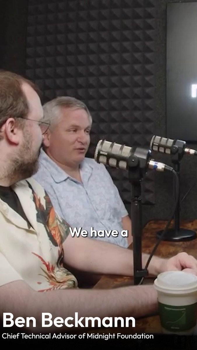 The image shows two men sitting at a table with microphones, suggesting they are participating in an interview or podcast recording session. One man has a cup of coffee next to him while the other man is wearing glasses and looking towards something off-camera. The caption reads "We have a".
