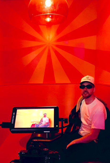 A man wearing sunglasses and a light cap sitting next to a camera monitor displaying a shot of himself, set against a vibrant red room with a radiating sunburst pattern on the wall.