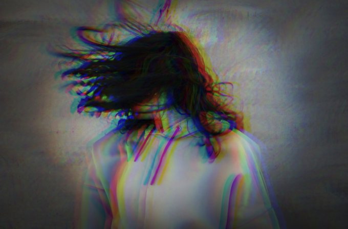 A figure with long flowing hair is shown from behind in a glitch-art style image featuring chromatic aberration with red, blue, and green color distortions against a textured background.