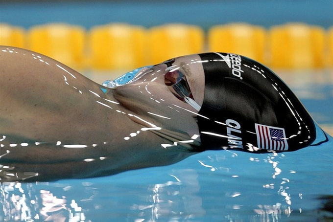 A swimmer wearing a black 'arena' swim cap with an American flag and goggles performs the backstroke in a pool, with yellow lane dividers visible in the background.