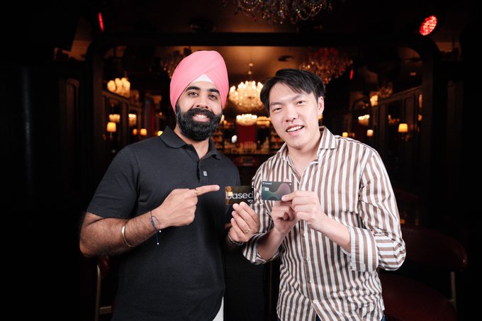 Two men pose at an upscale venue, with the man on the left wearing a pink turban and black polo pointing at a card, while the man on the right in a striped shirt holds up a "Based One" branded card, referencing their collaboration on the @BasedOneX project.