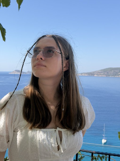 A young woman with long hair and sunglasses in a white dress stands in front of a bright blue ocean with distant hills and a sailboat visible.
