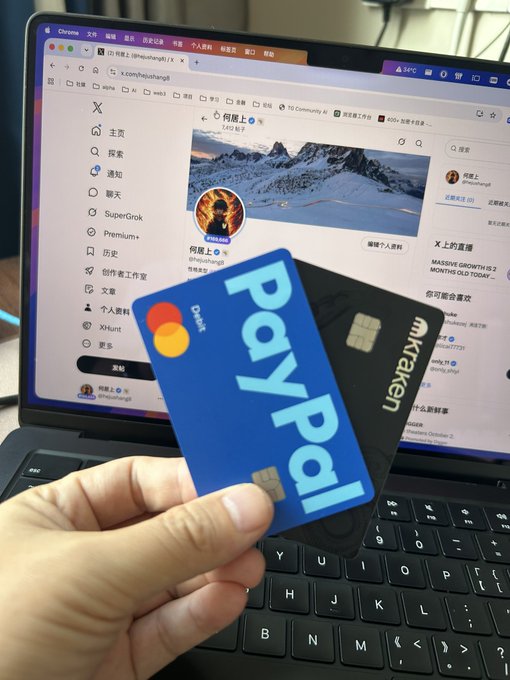 A hand holds up a blue PayPal Debit card and a black Kraken card in front of a laptop displaying X (formerly Twitter), where the user "何爱上" (@hezuoshangh) appears to have shared their experience of receiving both cards after nearly a month-long process, ready to test PayPal's fee-free cash withdrawals and real-time exchange rates.