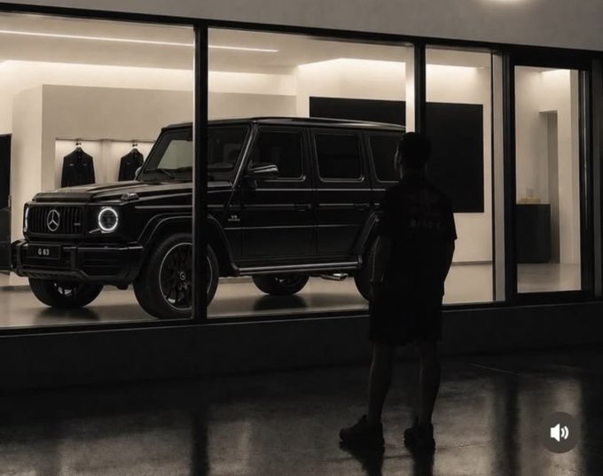 A person stands in a dark room looking through a large window at a black Mercedes-Benz G-Class SUV displayed in a well-lit showroom.