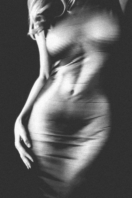 A black and white artistic photograph showing a pregnant woman's silhouette wearing a striped dress, with her hand gently cradling her belly, creating a soft contrast against the dark background.