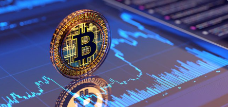 A close-up image of a physical Bitcoin token standing upright in front of a digital screen displaying a blue financial candlestick chart.