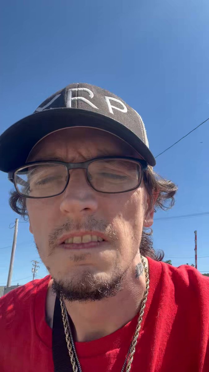 The image features a man wearing glasses and a red shirt with "XRP" written on his hat. He appears to be looking off into the distance or at something out of frame, possibly lost in thought. The background shows an open sky, suggesting that he might be outdoors during the day.