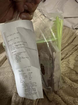 A hand holding up a printed receipt and a clear plastic bag containing what appears to be a packaged food product with green and brown elements, resting against a textured beige surface.