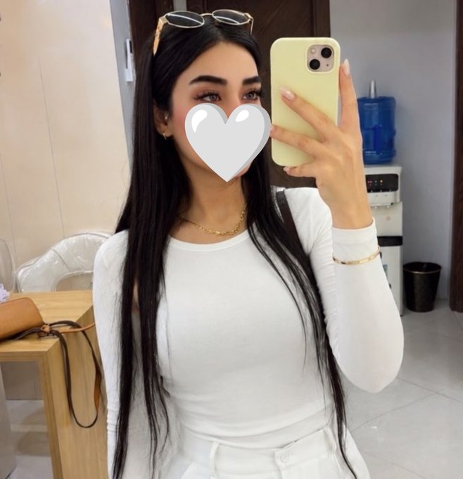 A woman with long dark hair takes a mirror selfie while wearing a white long-sleeved top, with a white heart covering her face and sunglasses perched on her head.
