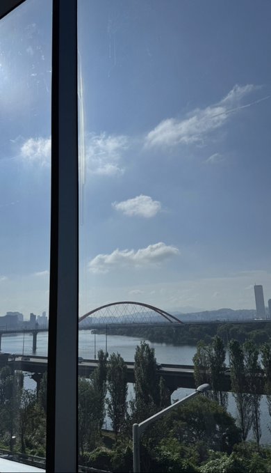 A scenic view through a window shows a river, an arched bridge, distant city buildings, and a blue sky with scattered clouds.