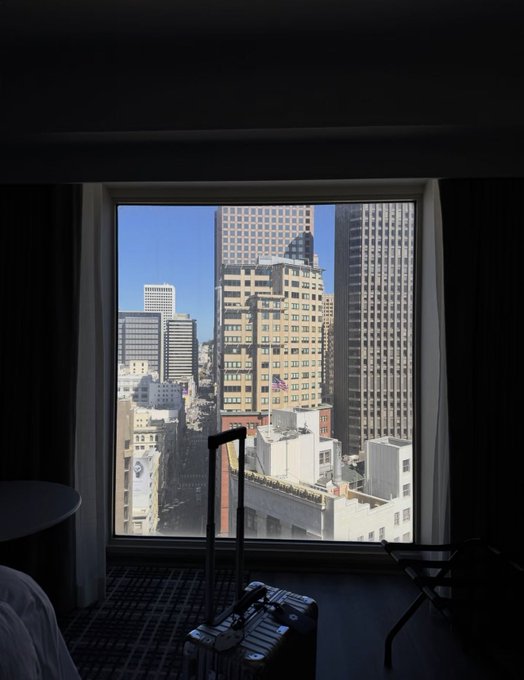 A photograph taken from inside a dim hotel room looking out a large window at a sunny cityscape with tall skyscrapers and a street below.