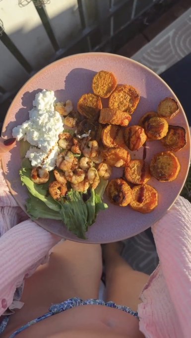 An overhead view shows a pink bowl filled with roasted sweet potatoes, shrimp, avocado, and a dollop of white cheese or cream, held over someone's lap in sunlight.