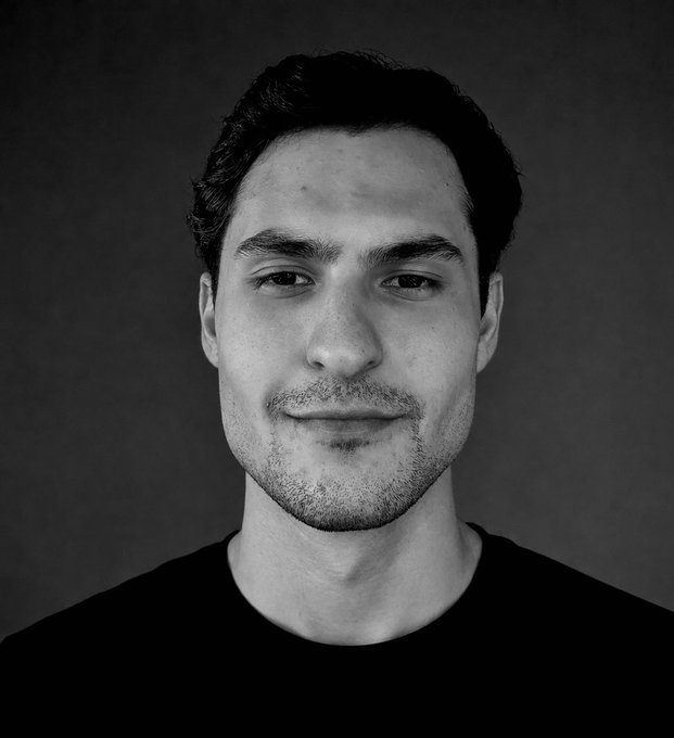 A black-and-white headshot of a young man with short dark hair and light stubble, wearing a black t-shirt, offering a subtle smile against a dark background—pairing with his post as a personal introduction while he seeks his next marketing and socials role in the Solana ecosystem.