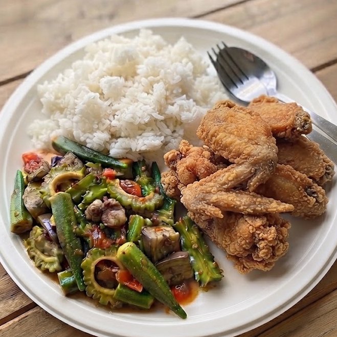 A white plate containing a serving of white rice, stir-fried bitter melon with eggplant, tomatoes, and okra, alongside several pieces of fried chicken, with a fork and spoon resting on the plate.
