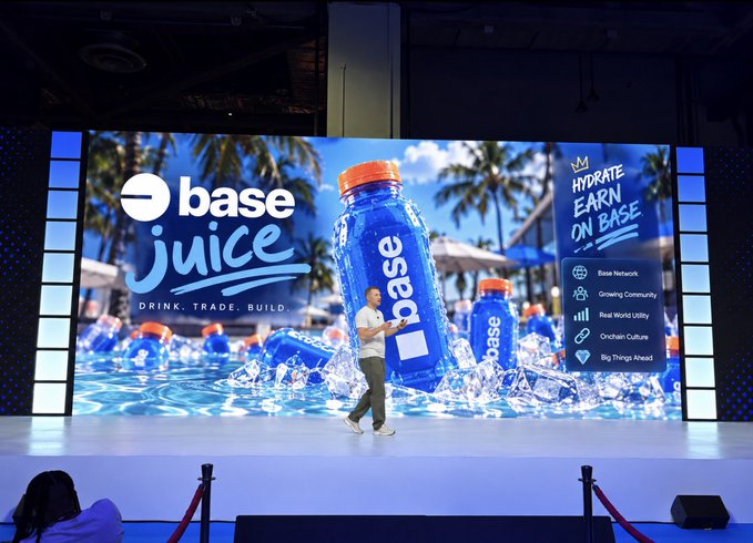 A man walks across a stage presenting 'base juice' (Drink. Trade. Build.) on a large screen displaying a product bottle, with text reading 'HYDRATE EARN ON BASE' and features like 'Base Network,' 'Growing Community,' 'Real World Utility,' 'Onchain Culture,' and 'Big Things Ahead.'