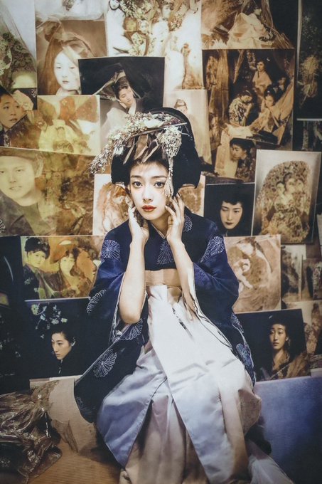 A young woman in an elaborate traditional dark blue and white Japanese kimono with ornate hair ornaments poses in front of a wall covered with artistic portrait photographs.