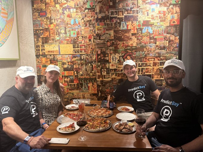 Four team members wearing "PredictBay" branded shirts and hats gather around a table filled with pizzas and wine glasses in front of a collage-covered wall, enjoying a team dinner ahead of the Sui Network event.