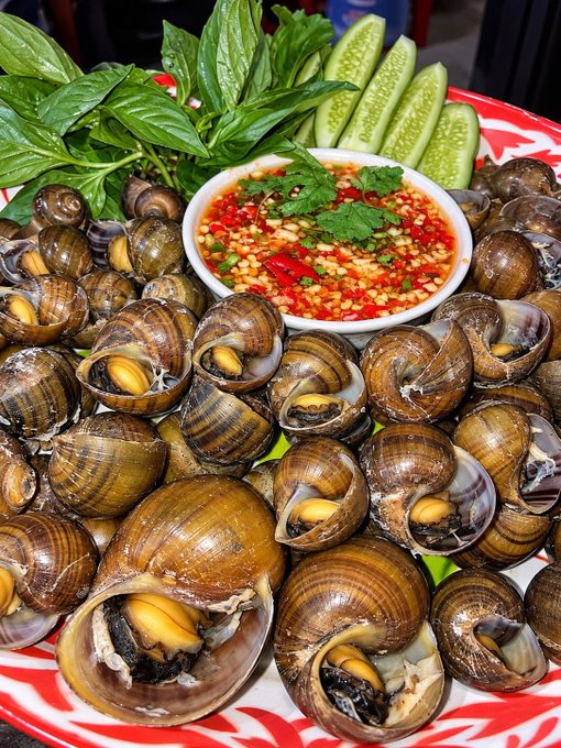 A plate of cooked snails served with fresh Thai basil, cucumber slices, and a bowl of spicy chili dipping sauce with cilantro.