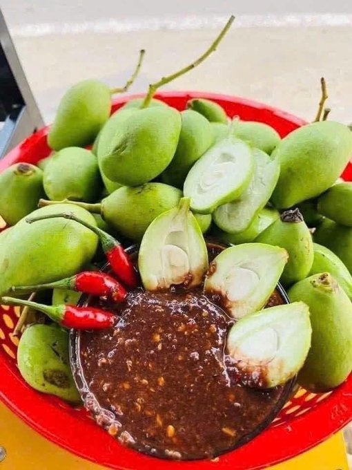 A red basket filled with green mangoes, some sliced open to reveal the flesh, served with a bowl of dark chili dipping sauce and fresh red chili peppers.