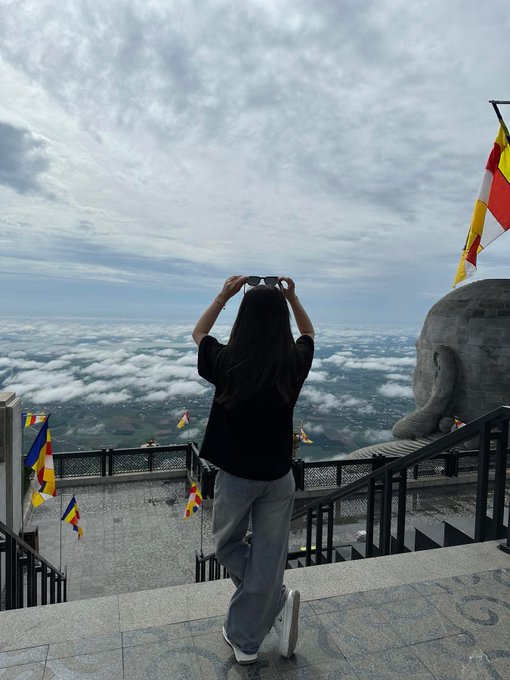 The image features a woman standing at the top of stairs, looking out over a beautiful view of the city below. She appears to be wearing sunglasses as she enjoys her time outdoors. The photo has been shared on social media with the caption "Chuyện mà bản thân phải quá ……. thiệt thòi !!! đầu tuần đã k zui rồi @wallchain @quipnetwork @sleepagotchi". The woman's sunglasses and the captivating view of the city create a visually appealing scene.