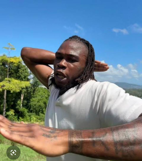 A muscular man with dreadlocks and a beard, wearing a white t-shirt and sporting arm tattoos, stretching his arms behind his head outdoors with green trees in the background.