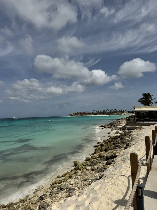 A scenic tropical beach with turquoise water, white sand, scattered rocks, and palm trees under a partly cloudy blue sky.