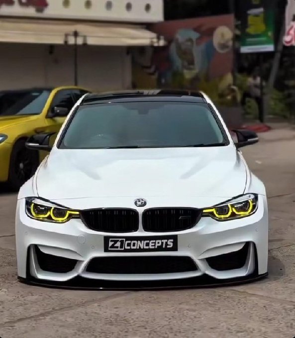 A white BMW car with yellow accent headlights and a '4 CONCEPTS' logo on the front bumper, parked in front of a graffiti-covered wall.