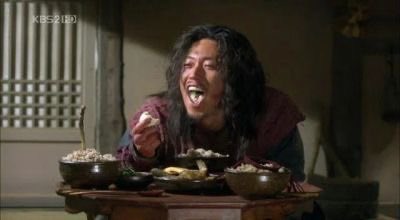 A person with long dark hair, wearing a dark red garment, laughs joyfully while eating from bowls on a wooden table, with a KBS2 HD logo visible in the upper left corner.