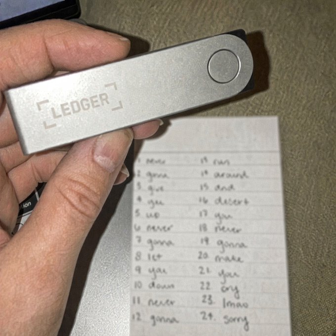 A hand displays a silver Ledger hardware wallet above a handwritten list of 24 recovery seed words, including "scare," "cheap," "child," "actual," reinforcing the post's message about securely storing recovery phrases for a newly set up crypto wallet.