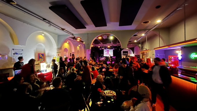 The image shows a large group of people gathered at an event, possibly a music festival or a social gathering. The crowd consists of many individuals standing around and interacting with each other. There are also some chairs placed throughout the scene, indicating that there might be seating arrangements for attendees. The atmosphere appears to be lively and engaging as people enjoy themselves in this public space.