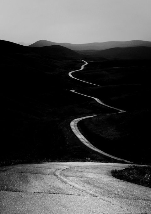 A winding mountain road descends through shadowy, silhouetted hills beneath a hazy sky, evoking a quiet, atmospheric sense of journey and solitude. The moody monochromatic tones reflect the artwork's title "Twilight," a 15-edition Tezos NFT.