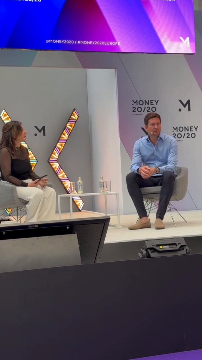 In the image, a man and woman are sitting at a table with a microphone, likely participating in an interview or panel discussion. The backdrop features a large screen displaying text related to cryptocurrency and financial institutions. The main focus of the image seems to be on the conversation between the two individuals rather than the content displayed on the screen.