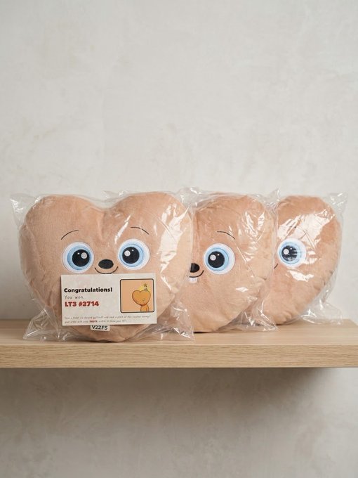 Heart-shaped peach-colored plush toys packaged in plastic on a wooden shelf, with the front one displaying a 'Congratulations! YOU WON' sticker with code LT3 83714.