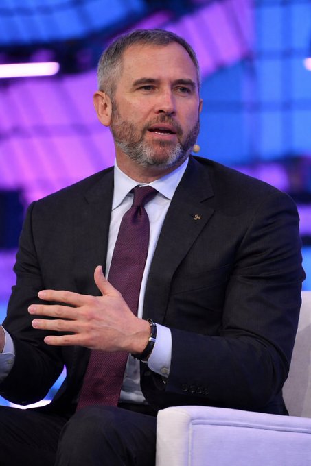 The image features a man dressed in a suit, sitting down with his hands together while wearing a purple tie. The background of the image shows a blue wall with white stripes, which appears to be part of an event or presentation setting. There is also text present that reads "For years they said Ripple only makes money by selling XRP." This suggests that the man in the suit might be discussing or talking about Ripple cryptocurrency and its financial implications during this event.