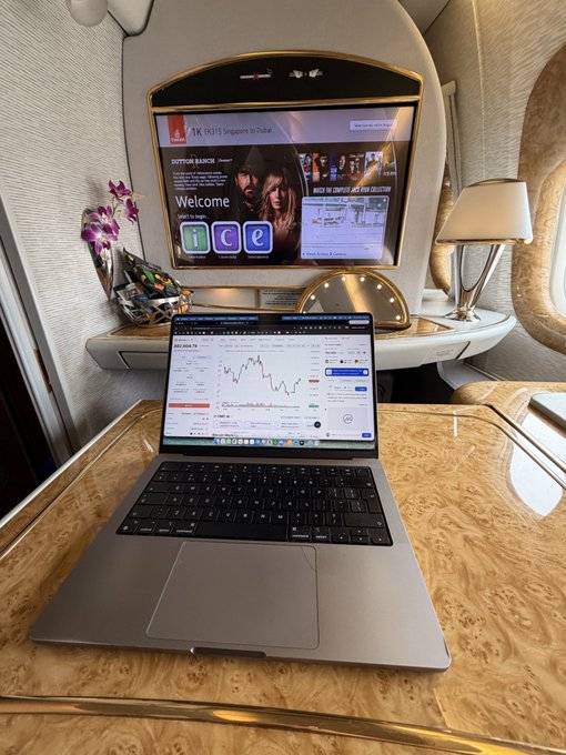 A MacBook displaying a stock chart for Singapore Airlines sits on a wood-grain table in what appears to be an Emirates first-class suite, with the in-flight entertainment screen behind showing a "1K" Emirates Skywards welcome message and a "Singapore to Dubai" route header. The setup captures a productive travel moment, fitting perfectly with the return journey home to Dubai.