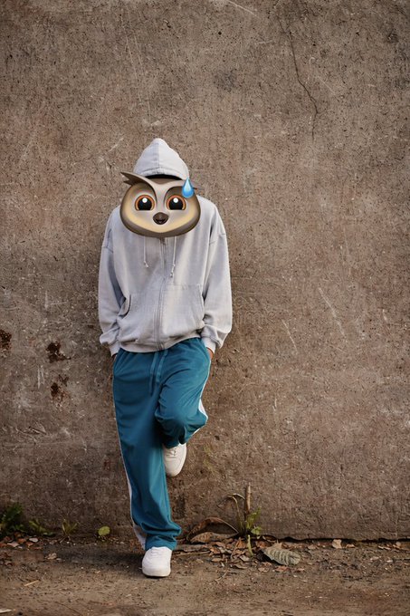 A person in a gray hoodie and teal pants leaning against a weathered concrete wall, with a cartoon animal emoji (a sweaty seal/otter face) covering their actual face.