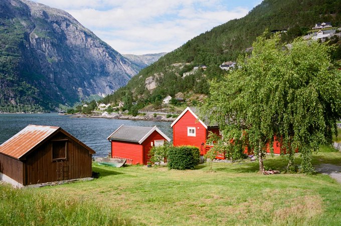 A scenic landscape features traditional red wooden cabins beside a fjord, surrounded by lush green grass, birch trees, and towering mountains under a partly cloudy sky.