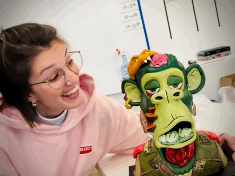 The image features a woman wearing glasses and a pink sweatshirt sitting next to a green monkey statue with a face painted on its head that appears to be smiling at her. The monkey's arms are raised, giving it an impression of being ready for action or play. The background shows a whiteboard filled with writing, suggesting the setting might be a classroom or office environment where the woman and the monkey statue could be part of an educational or creative project.
