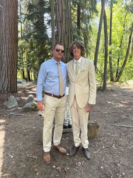 Two men in suits stand together in a wooded outdoor setting, with one in a light blue shirt and the other in a beige suit with a tie.