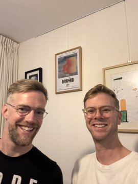 A selfie of two smiling men with glasses standing indoors in front of a white wall decorated with framed artwork.