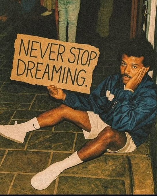 Vintage photo of Lionel Richie sitting on a sidewalk wearing a blue jacket and white shorts, holding a cardboard sign that reads 'NEVER STOP DREAMING.'