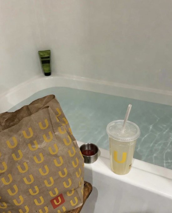 A brown paper bag printed with repeating yellow "U" logos sits next to a bathtub filled with water, alongside a clear plastic cup with a straw also bearing the "U" logo.