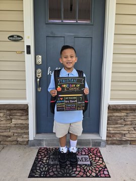 A young boy stands in front of a blue door holding a 'First Day of School' sign, wearing a backpack and a light blue shirt with shorts.