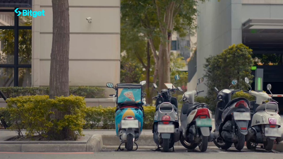 The image features a row of motorcycles parked next to each other, with one motorcycle having a blue paint job that says "bitget" on it. The motorcycles are lined up along a curb near some bushes, creating an interesting scene for viewers. In the background, there is a building and trees, adding more context to the location where these motorcycles are parked. The image also includes a caption about Bitget's Blockchain4Youth project, which could be related to the motorcycles or simply a random piece of information in the photo.