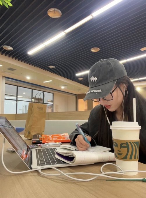 A young woman wearing a 'brand' cap and glasses studies intently at a Starbucks, writing in a notebook next to her laptop with earphones in.