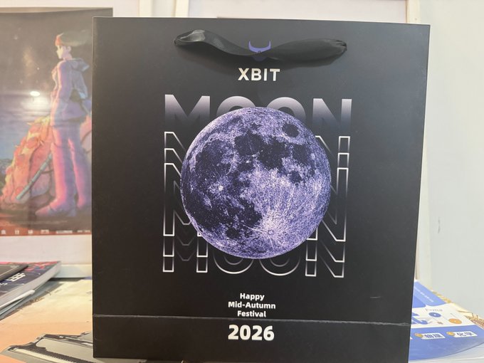 A black gift bag from 'XBIT' featuring a purple moon graphic with the text 'MOON' repeated and a label reading 'Happy Mid-Autumn Festival 2026'.