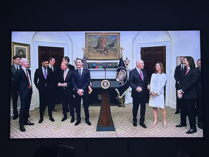 A live broadcast from the White House shows President Trump standing alongside an official group of advisors, including senior administration figures and a man in a light blue suit believed to be Brad Garlinghouse, gathered around the official podium prepared for an imminent announcement or major appointment.