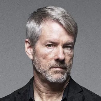 A distinguished-looking man with gray hair and a beard, wearing a dark blazer, poses against a neutral gray background. The accompanying post's provocative message about Bitcoin policy reform creates an ironic juxtaposition with the portrait's formal, headshot-style composition.