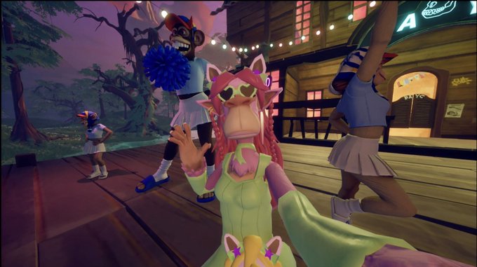 A 3D avatar with pink hair and heart-shaped sunglasses waves in a virtual environment with other avatars, including one with a blue pom-pom, on a wooden platform near a building lit with string lights.