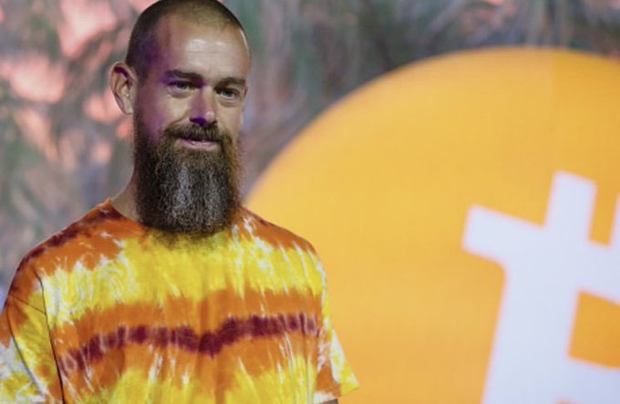 A bearded man in a yellow and orange tie-dye shirt stands in front of a backdrop featuring an orange Bitcoin-themed logo, embodying the post's message of humility and simplicity in the Bitcoin community.